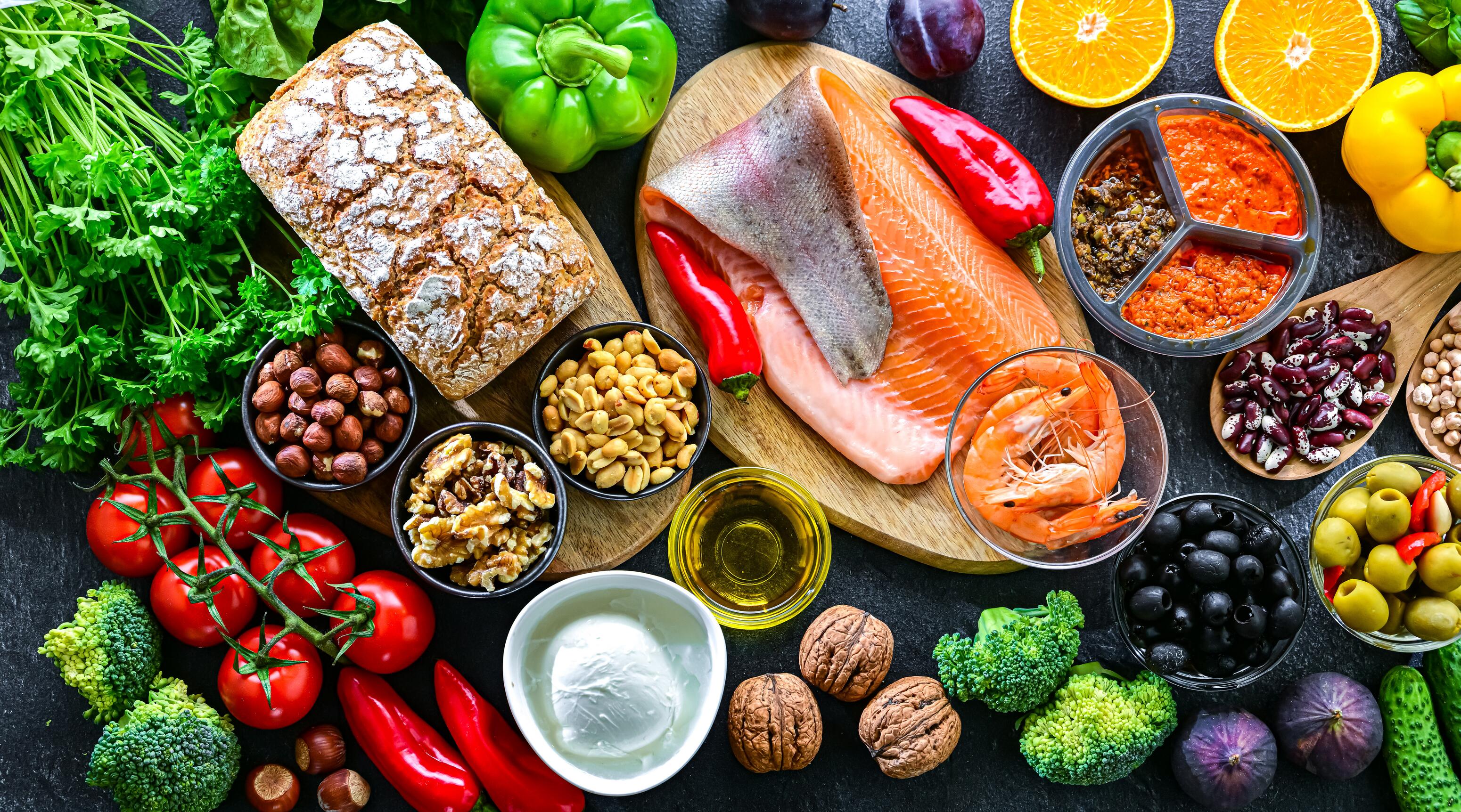 Auf einem Tisch sind zahlreiche Lebensmittel, wie Lachs, Tomaten, Oliven, Brokkoli, Nüsse, Paprika, Orangen und mehr zu sehen.